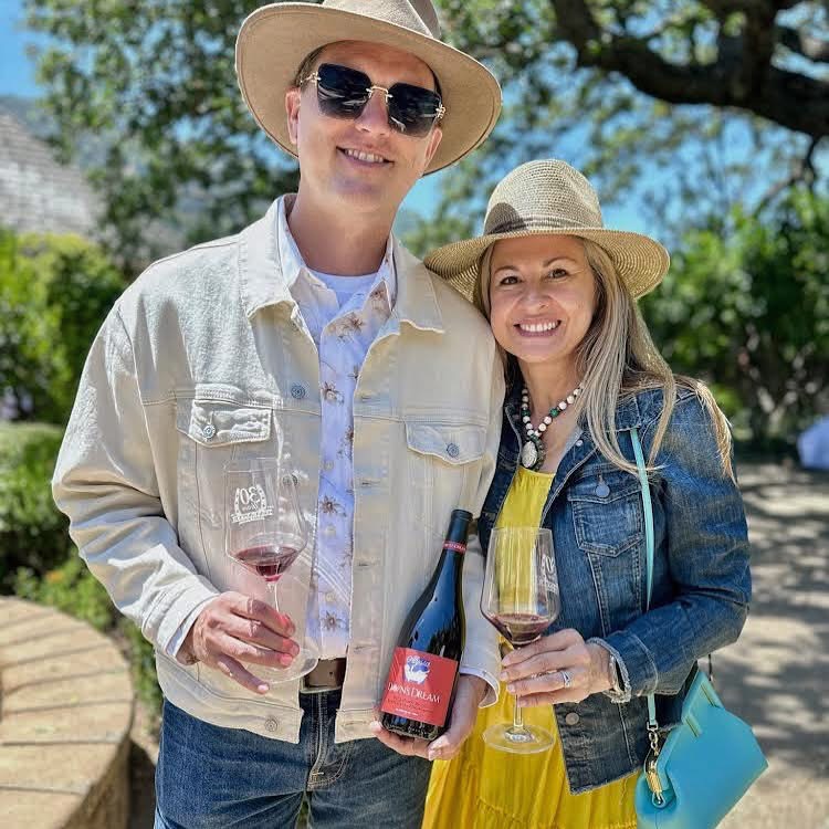 Smiling man and woman in hats hold wine glasses and a wine bottle outdoors on a sunny day.