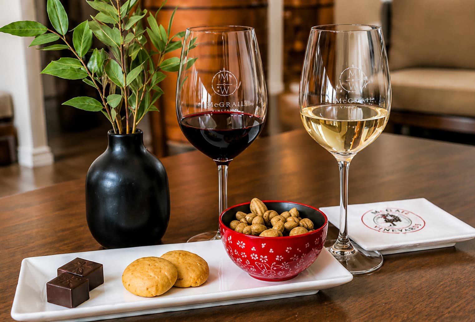 Two glasses of wine, a bowl of nuts, cookies, and chocolates on a table with a small black vase and napkins.