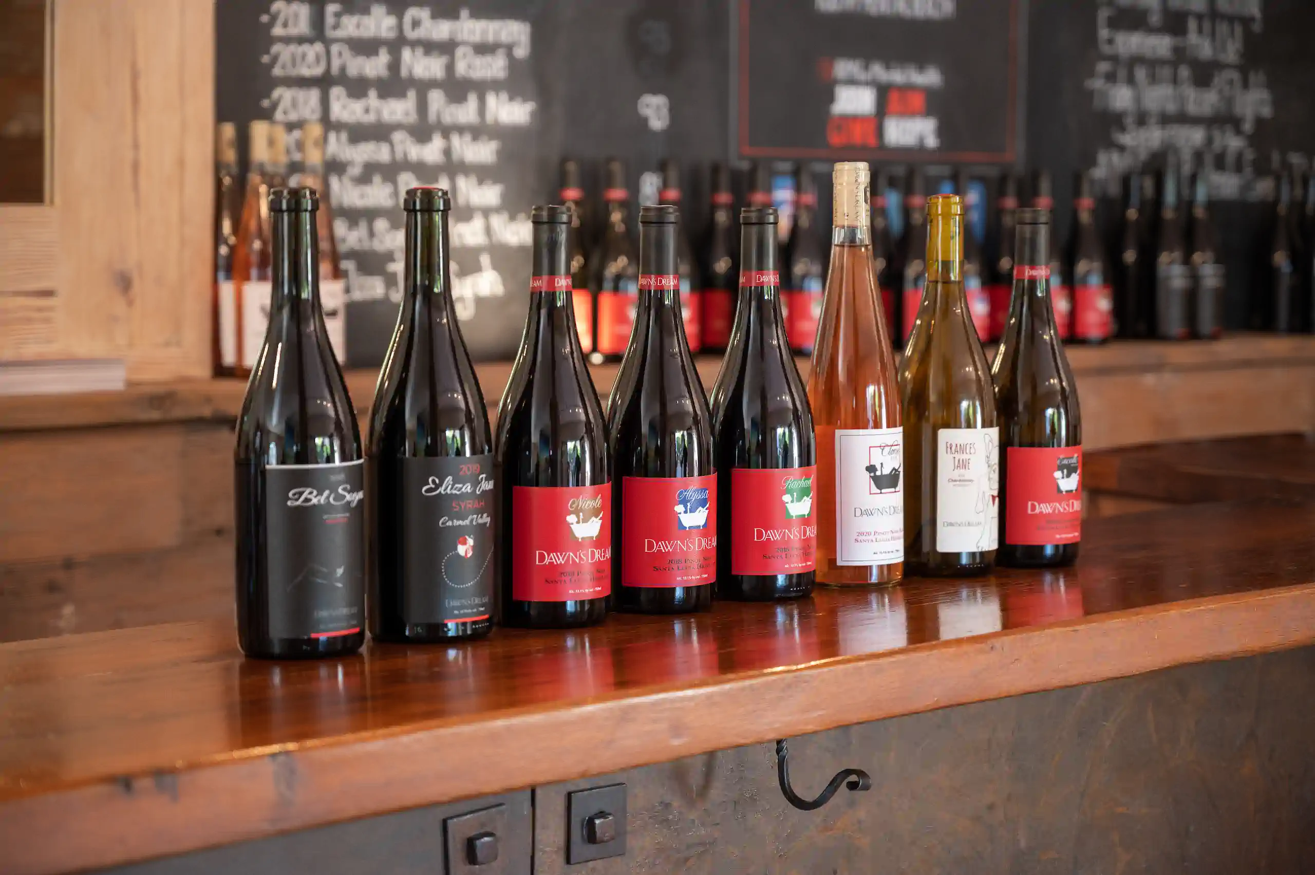 Nine bottles of wine lined up on a wooden counter, with more bottles and a chalkboard in the background.