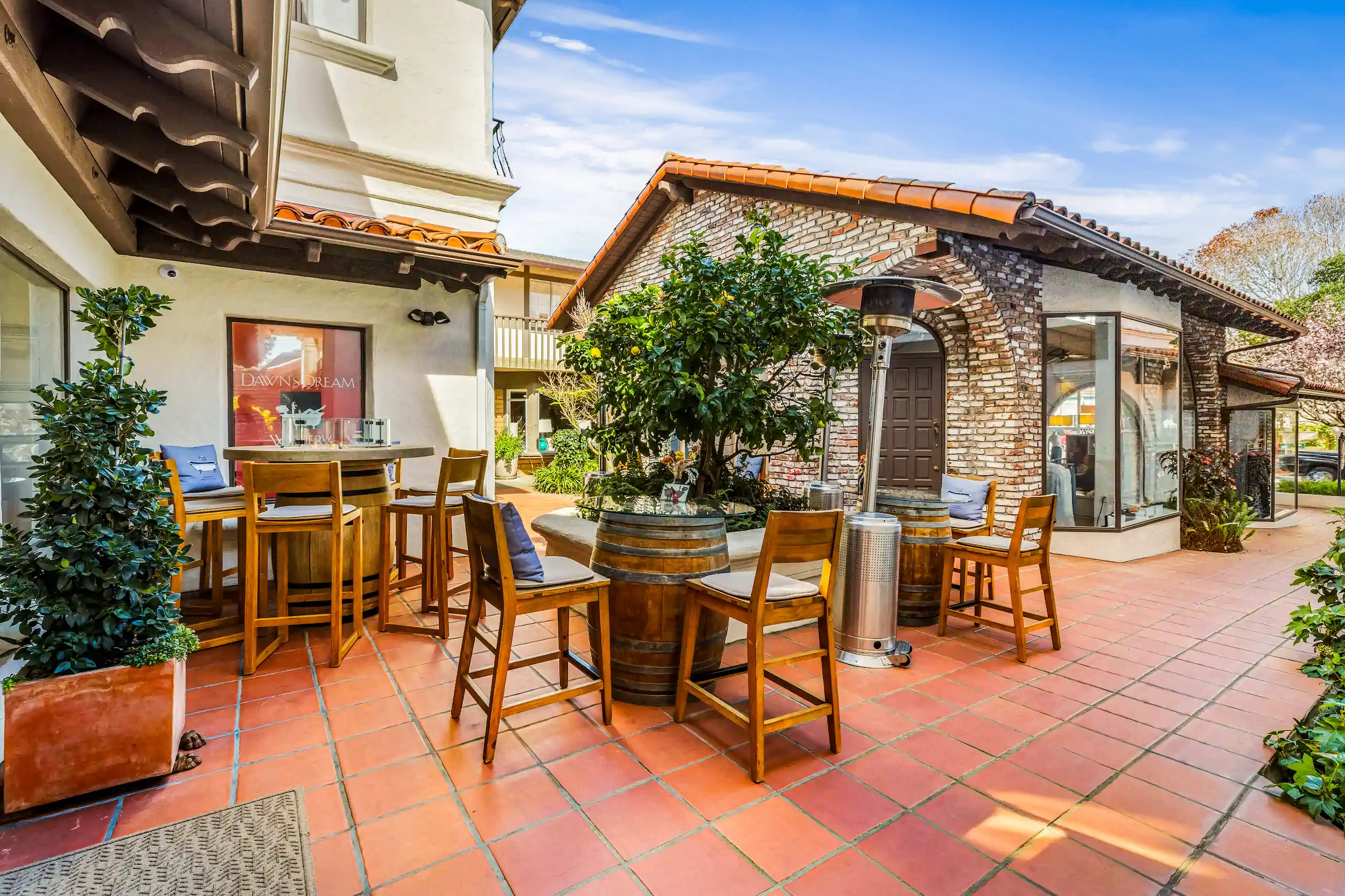 Sunny outdoor patio with wooden tables and chairs, potted plants, and brick buildings with terracotta roofs.
