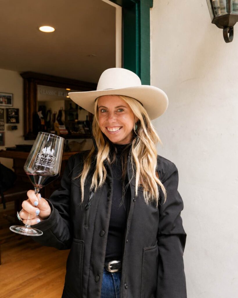 Woman in a hat smiles and holds a glass of red wine, standing in a doorway of a well-lit room.