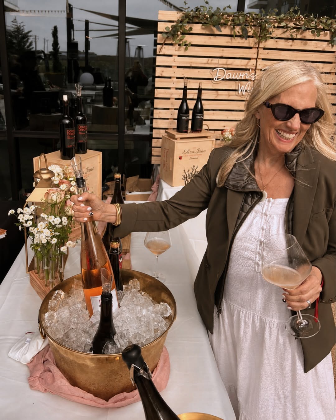 Woman in sunglasses smiles, holding wine, with bottles in ice bucket at an outdoor wine tasting event.
