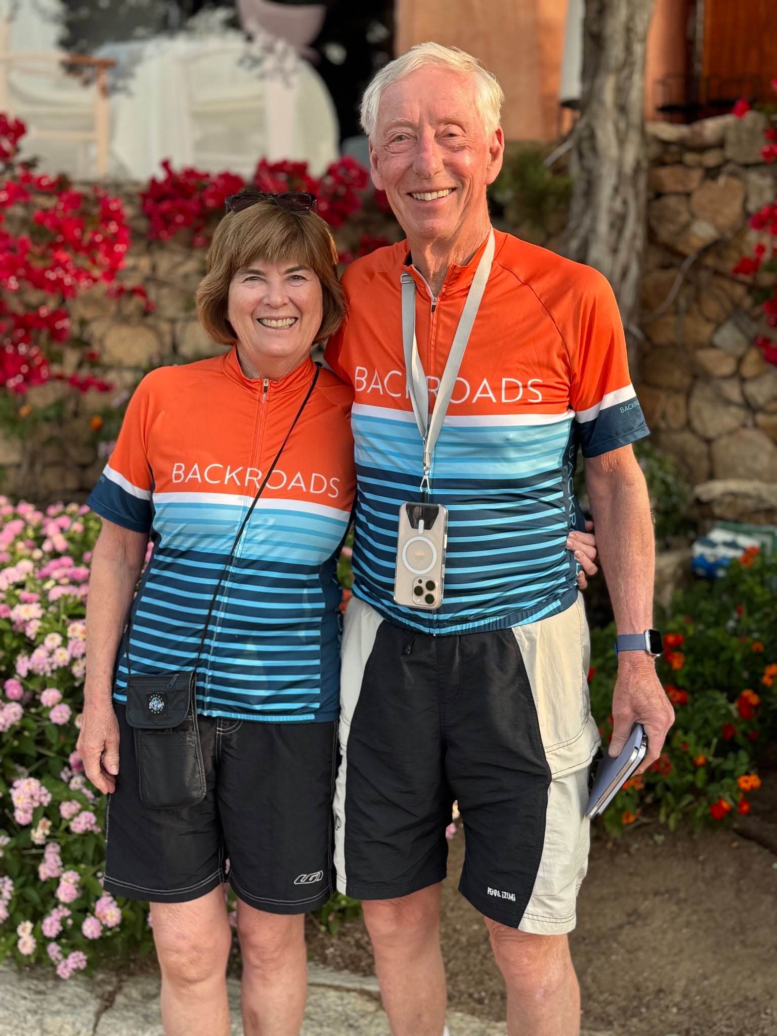 Two smiling people in matching cycling jerseys stand outside, surrounded by flowers and greenery.