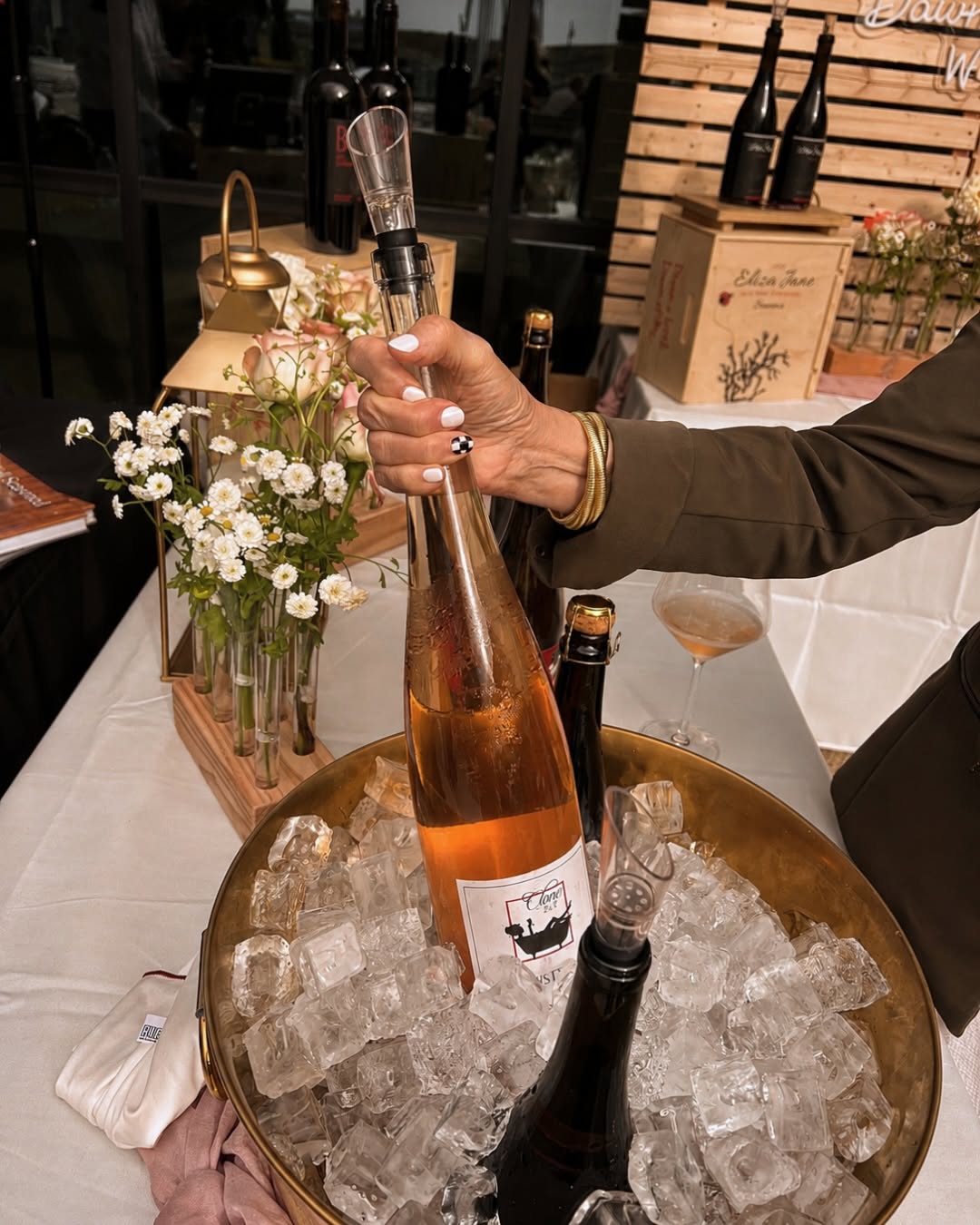 A hand holds a bottle of rosé wine in a gold ice bucket with flowers and bottles on a table.