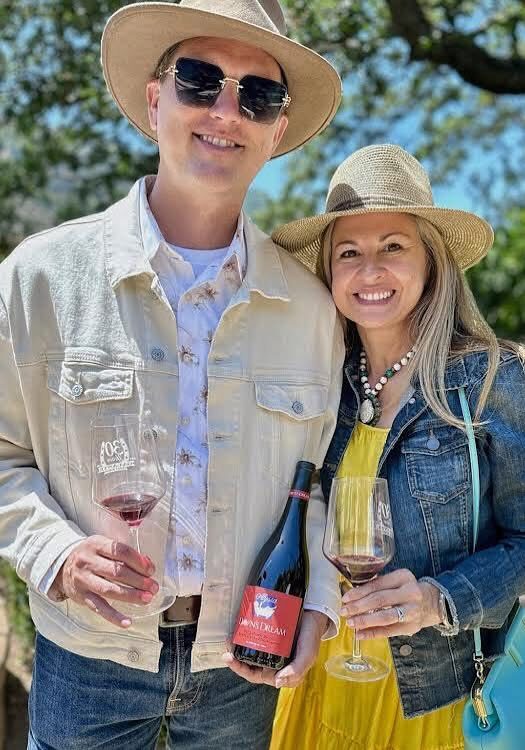 Smiling man and woman in hats hold wine glasses and a wine bottle outdoors on a sunny day.