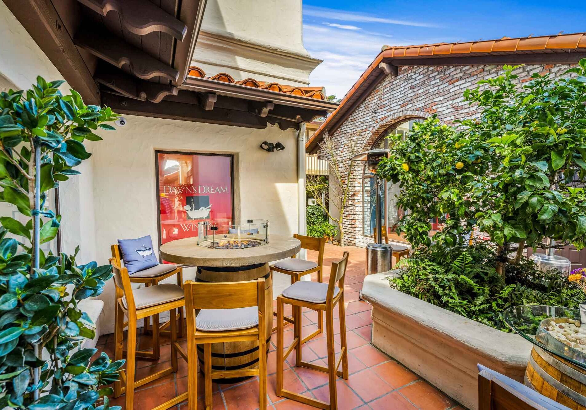 Outdoor patio with wooden chairs around a round table, surrounded by greenery and Spanish-style architecture.