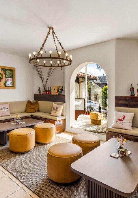 Bright lounge with beige sofas, round stools, wine bottles, and a large chandelier by an arched doorway.