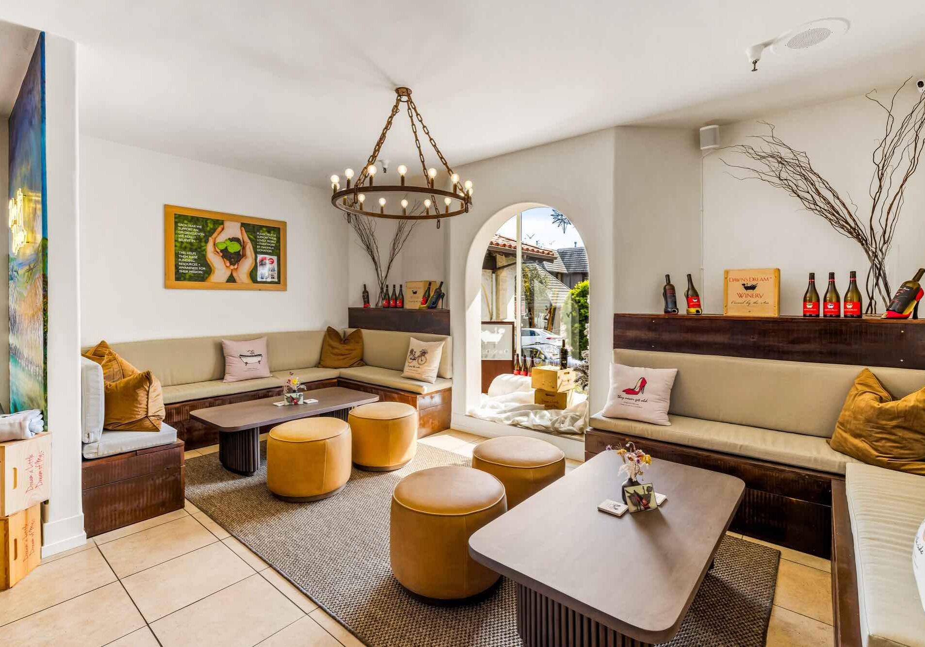 Bright lounge with beige sofas, round stools, wine bottles, and a large chandelier by an arched doorway.