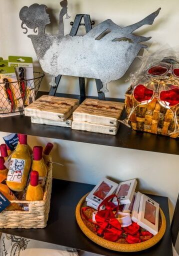 Shelf with wine bottles, coasters, red glasses, metal rooster art, and a hat on the side.
